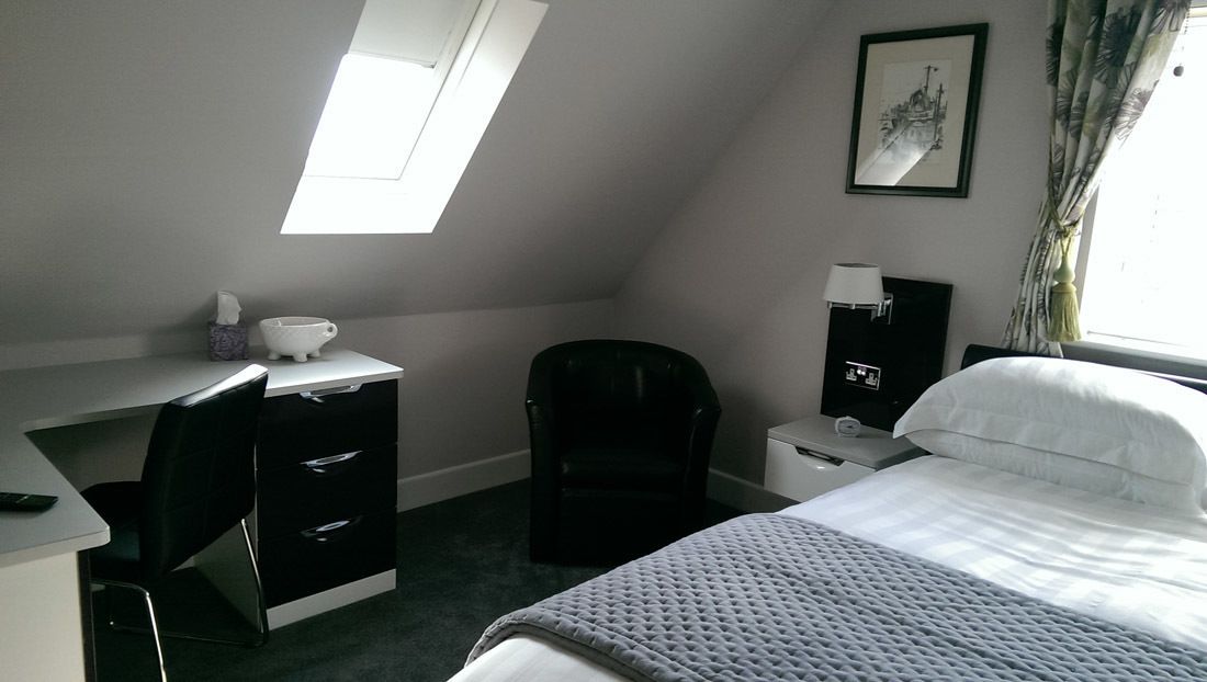 Una habitación de hotel pequeña con escritorio, silla, cama y una mesita de noche con lámpara.
