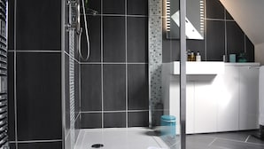 A modern bathroom with dark tiles, a glass-enclosed shower, a white sink, and a mirror. - Canford Crossing (Wimborne)