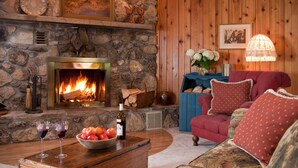 A cozy living room with a stone fireplace, a red armchair, and a wooden coffee table with wine glasses and a bowl of fruit. - Chanticleer Guest House (Sturgeon Bay)
