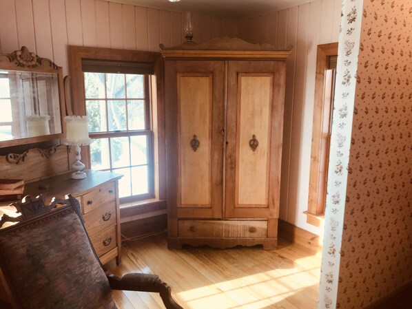 Una habitación con paneles de madera, un armario grande de madera, un escritorio con cajones, una silla y un ventanal con vistas a árboles.
