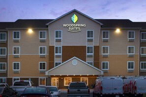 A multi-story hotel with a central entrance, multiple windows, and a parking lot with vehicles. - WoodSpring Suites Chicago Romeoville (Romeoville)