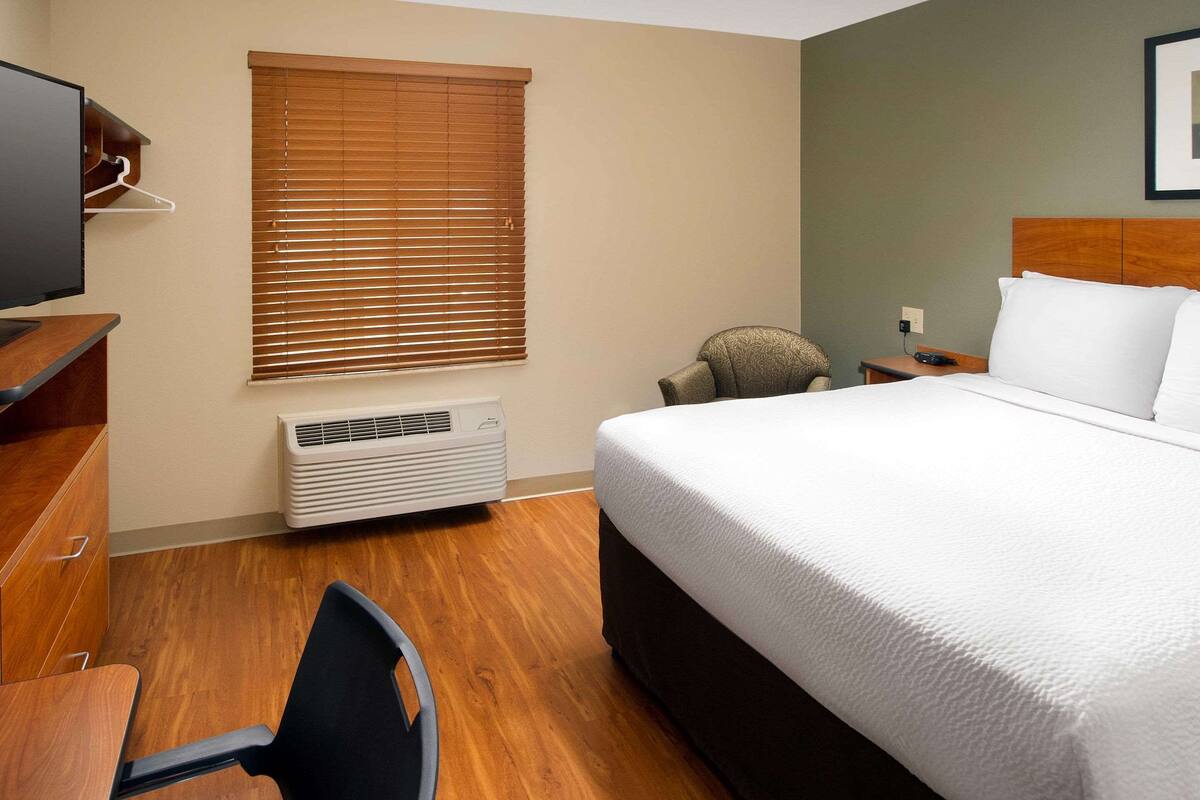 A hotel room with a bed, a desk, a chair, a TV, a heater, and a window with blinds.