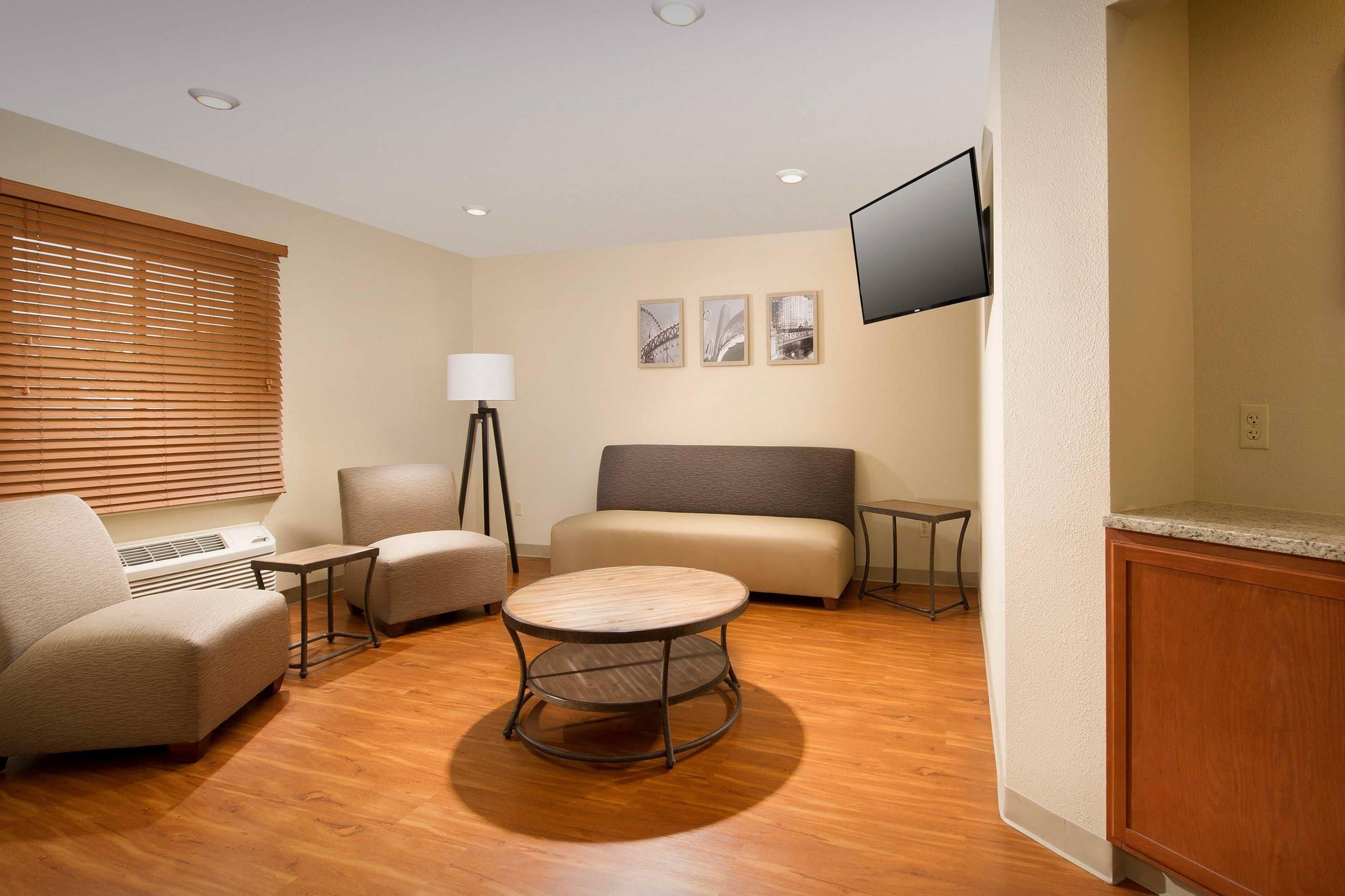 A hotel room with a sofa, two armchairs, a small table, a floor lamp, and a flat-screen TV mounted on the wall.