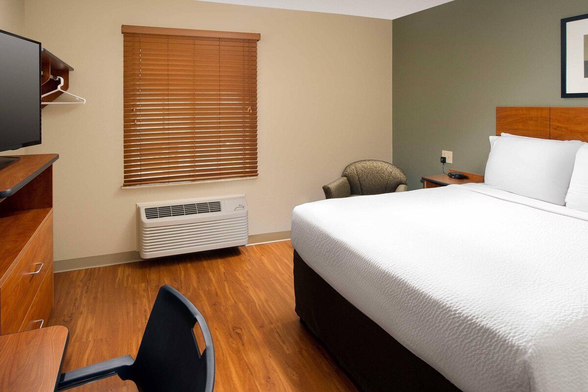 A hotel room with a bed, a desk, a chair, a window with blinds, and a wall-mounted air conditioner.