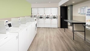 Una sala de bugaderia amb diverses rentadores i assecadores, una taula i una porta que dona a una altra habitació.