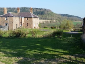 A stone building with a sloped roof, surrounded by a grassy area with a fence and trees.