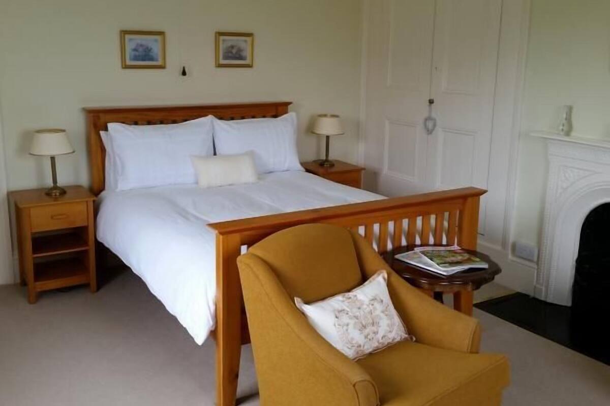 Un dormitorio ordenado con una cama de madera, dos mesitas de noche con lámparas, una silla y una chimenea.