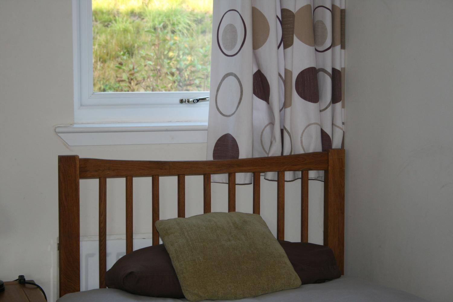 Un lit en bois avec une tête de lit, une fenêtre donnant sur un paysage verdoyant et un rideau orné d’un motif circulaire.