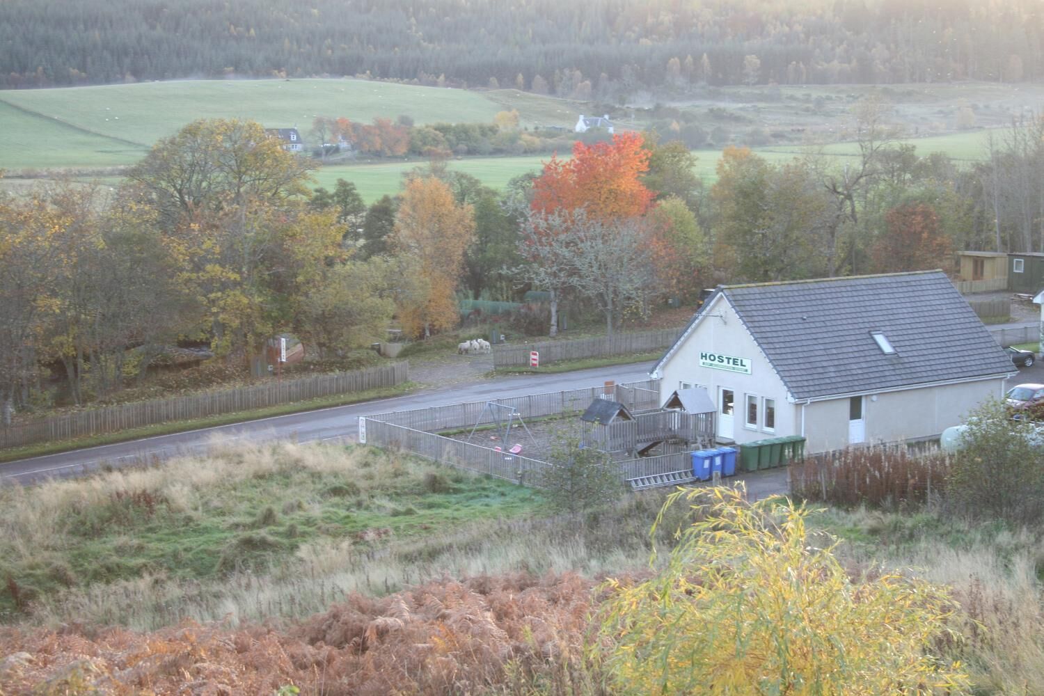 A hostel with a simple, functional design, located near a road with a fenced area and a field in the background.