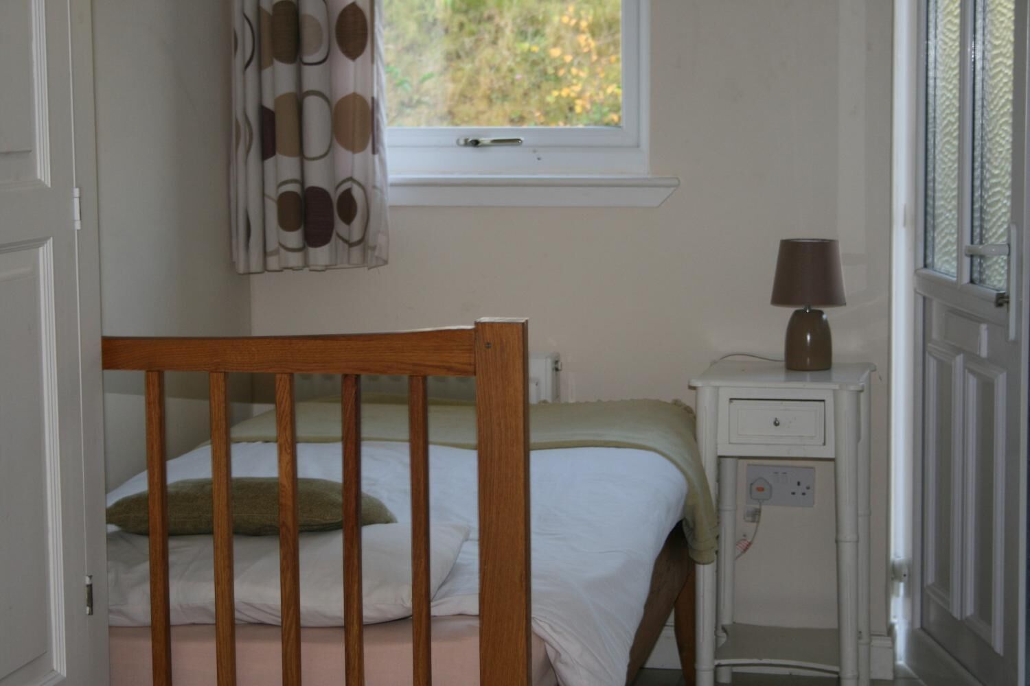 A room with a wooden crib, a bedside table with a lamp, a window with a curtain, and a wall-mounted electrical outlet.