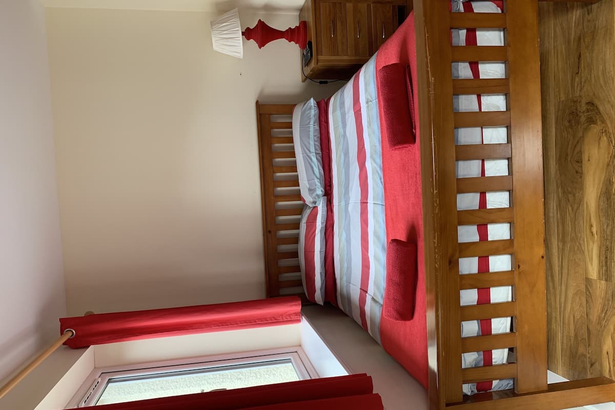 A small, compact room with a bunk bed, a window with a red blind, and a wooden headboard with a slatted design.
