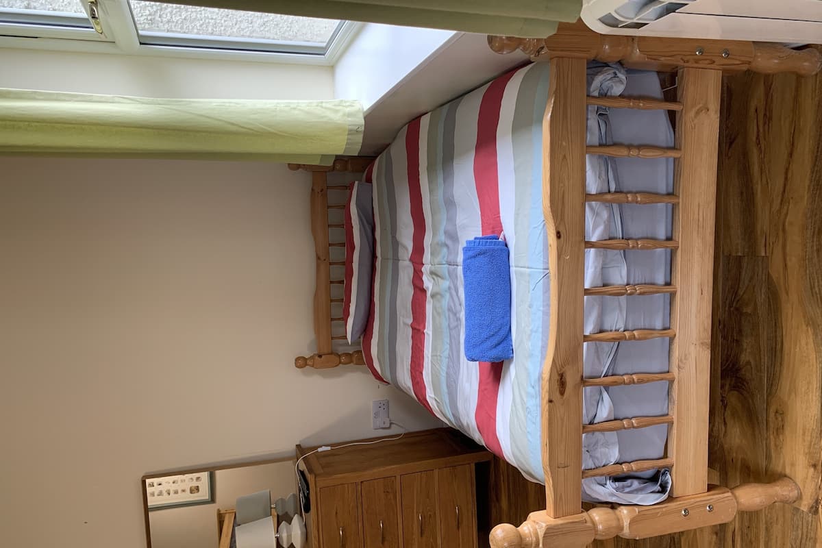 A single bed with a wooden frame, a striped bedspread, and a blue towel. There is a wooden wardrobe below the bed and a mirror on the wall.