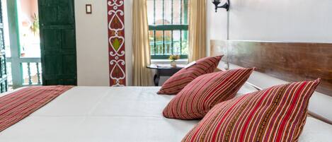 A hotel room with a large bed, striped pillows, a wooden headboard, a window with curtains, and a wall-mounted lamp.