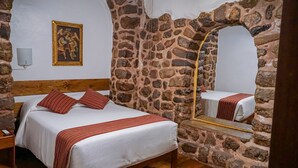Una habitación con pared de piedra, cama con ropa de cama blanca y almohadas a rayas rojas, cabecero de madera, un cuadro colgado en la pared y un espejo con marco de madera.