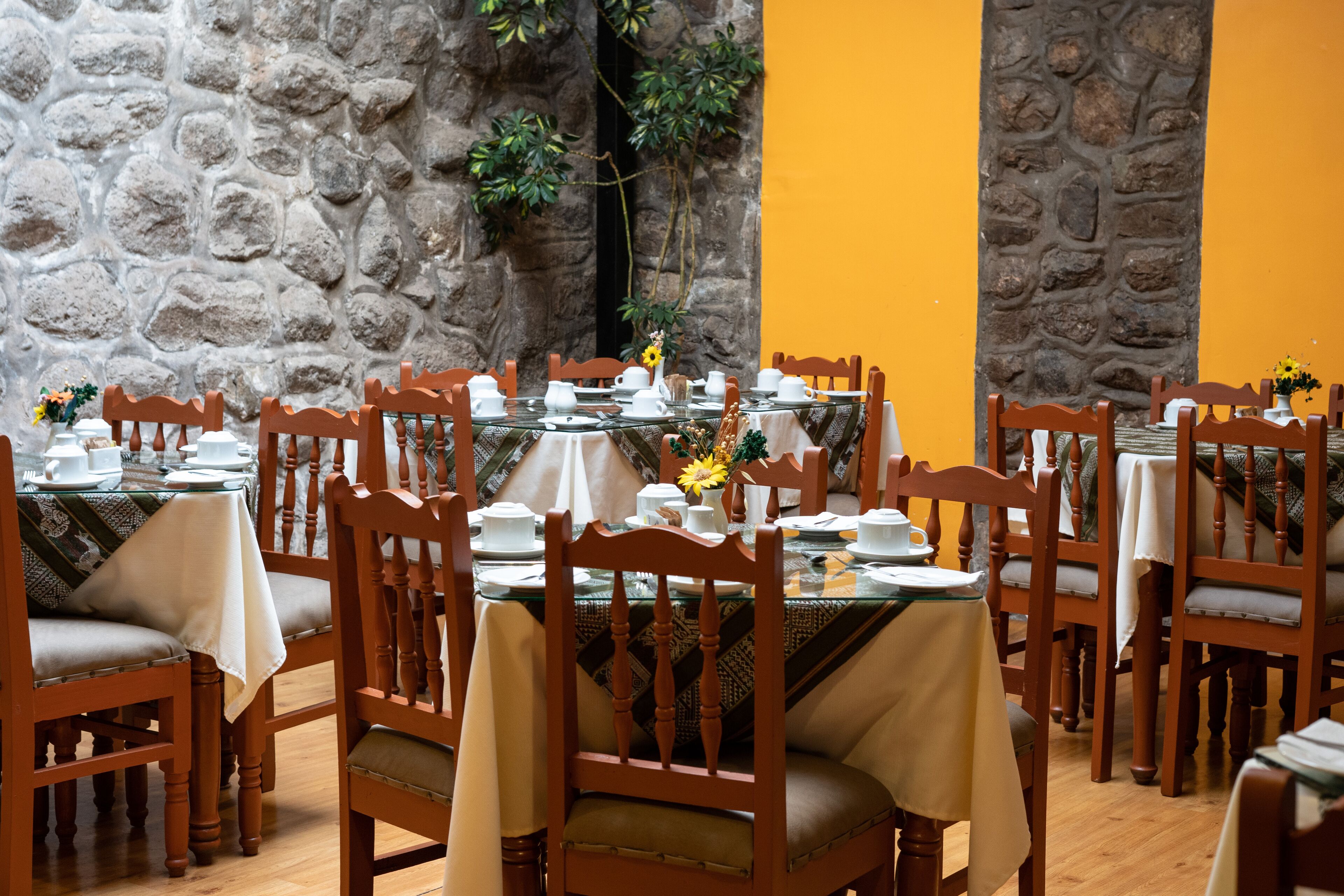 A dining area with multiple tables set for service, wooden chairs, and a stone wall backdrop.