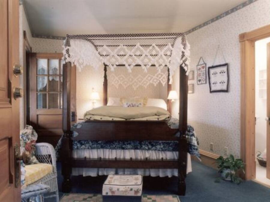 Una cama con dosel adornada con dosel de encaje, una mesita de noche, una silla y una pequeña mesita auxiliar.