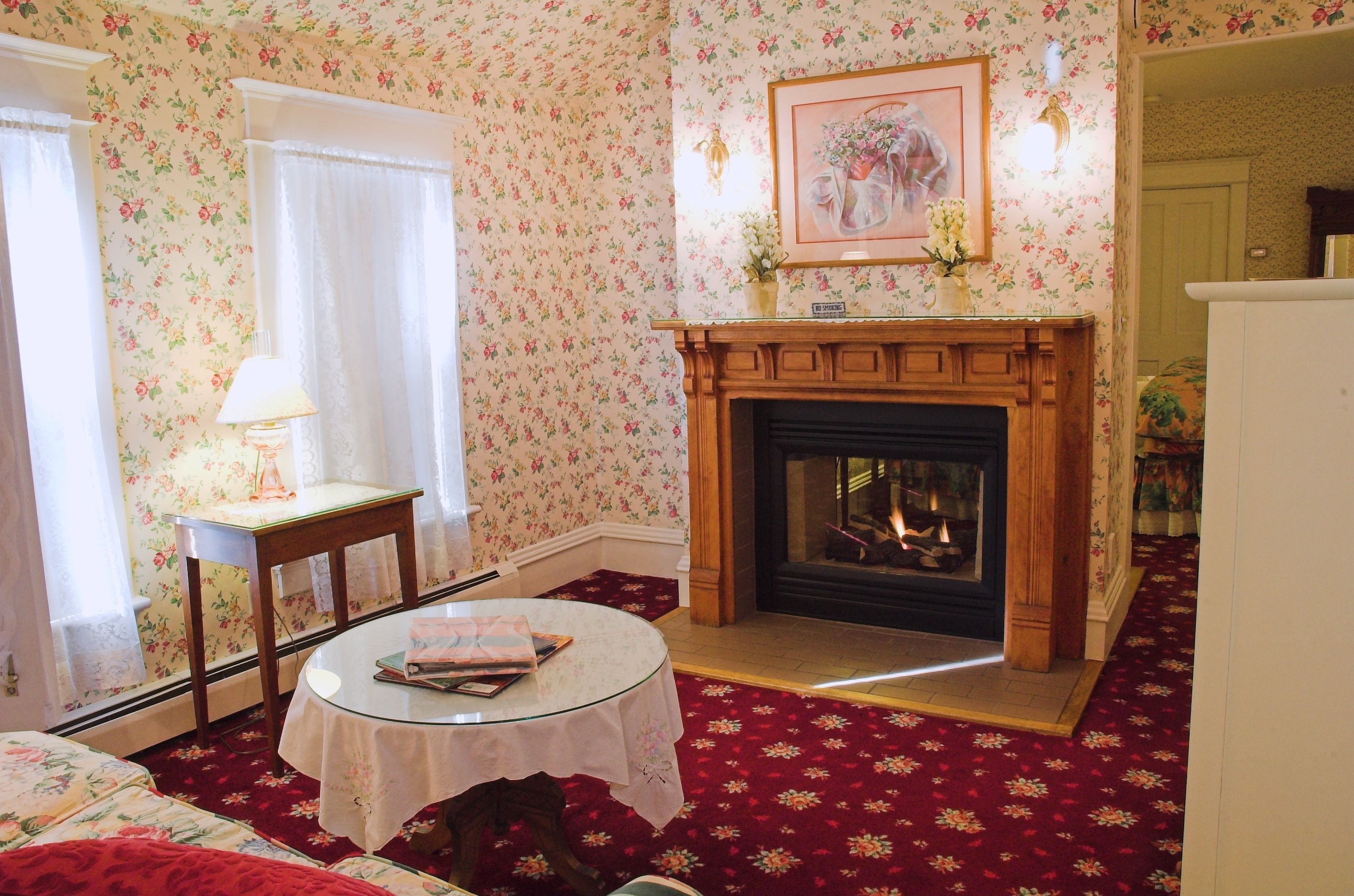 Una habitación con papel tapiz floral, una chimenea, una mesa redonda con un libro y una cama con ropa de cama floral.