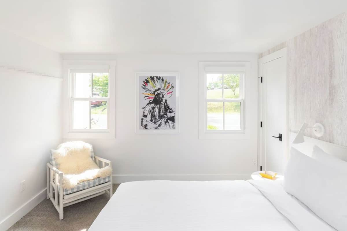 Un dormitorio luminoso y amplio, con una cama grande, dos ventanas, una silla y una obra de arte colgada en la pared.