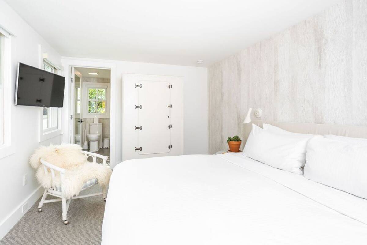 Un dormitorio con una cama grande, una silla blanca con una manta de piel, un televisor montado en la pared y una puerta que da a un baño.