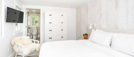 Un dormitorio con una cama grande, una silla blanca con una manta de piel, un televisor montado en la pared y una puerta que da a un baño.