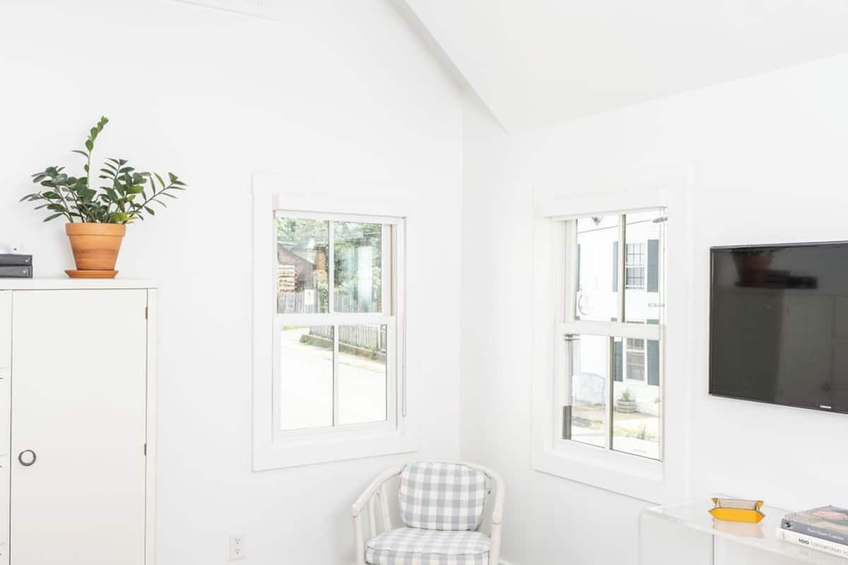 Una habitación luminosa y blanca con techo inclinado, un televisor montado en la pared, un ventanal pequeño y una silla con cojín de estampado cuadriculado.