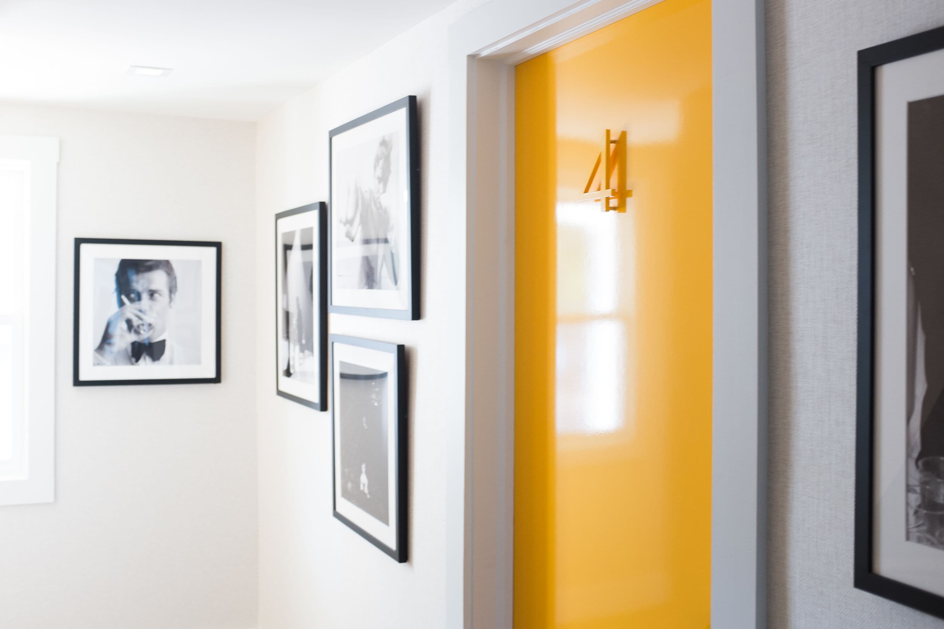 Un pasillo con fotografías enmarcadas en la pared, una puerta amarilla y un cuadro enmarcado a la derecha.