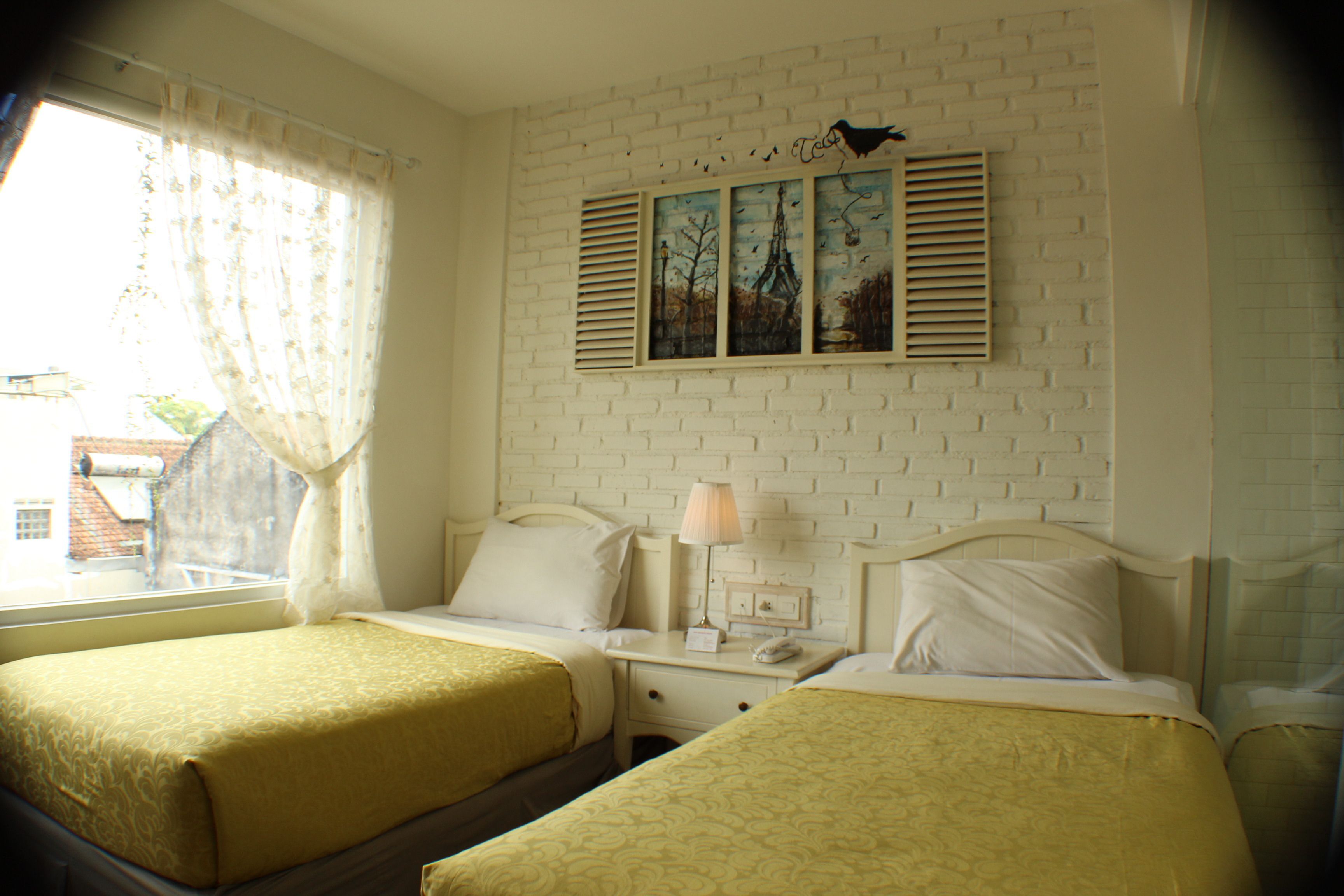 A hotel room with two beds, a window with curtains, a white brick wall, and a painting above the beds.