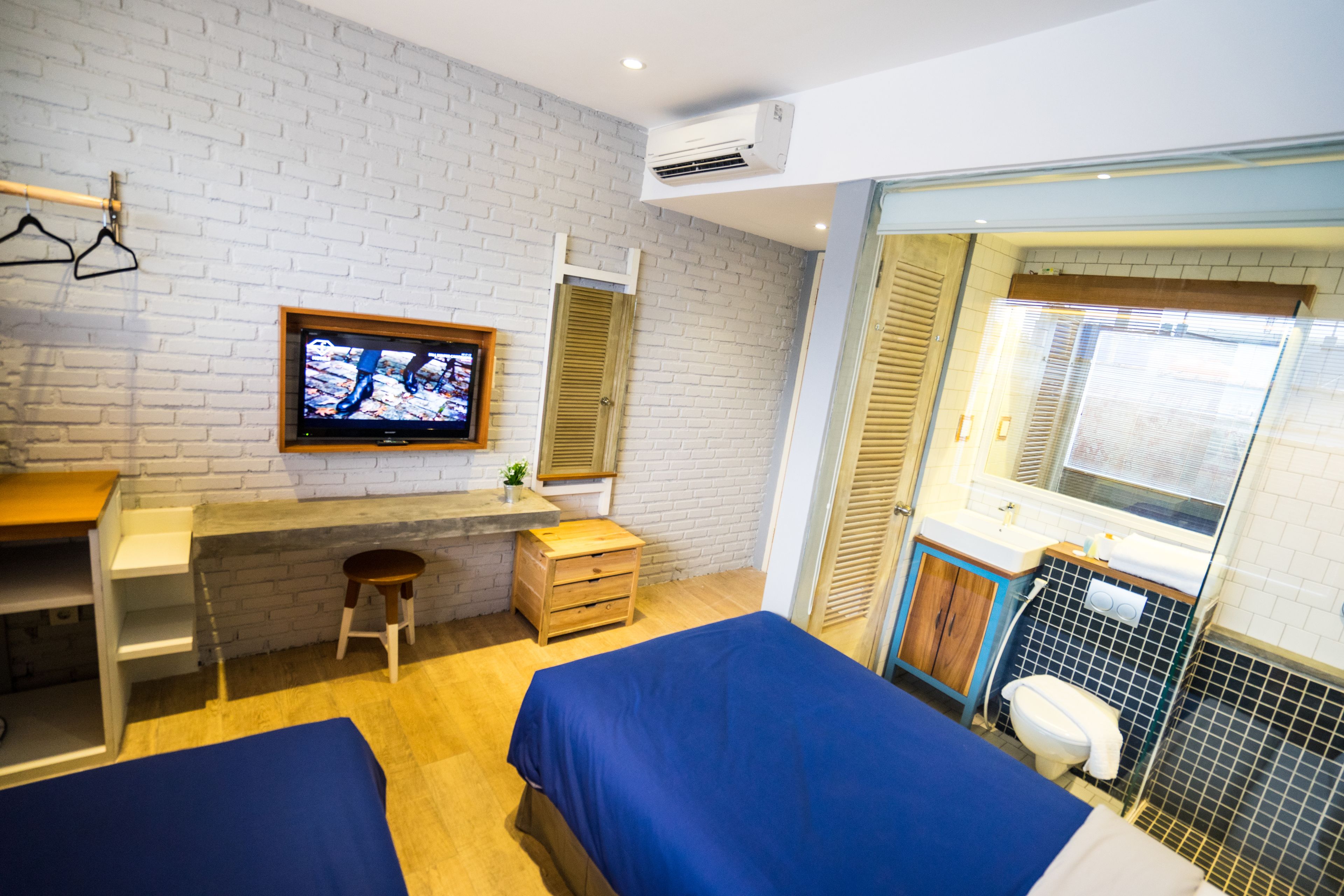 Habitación de hotel con pared de ladrillo blanco, escritorio de madera con televisor, dos camas, un taburete pequeño y un baño con ventana.