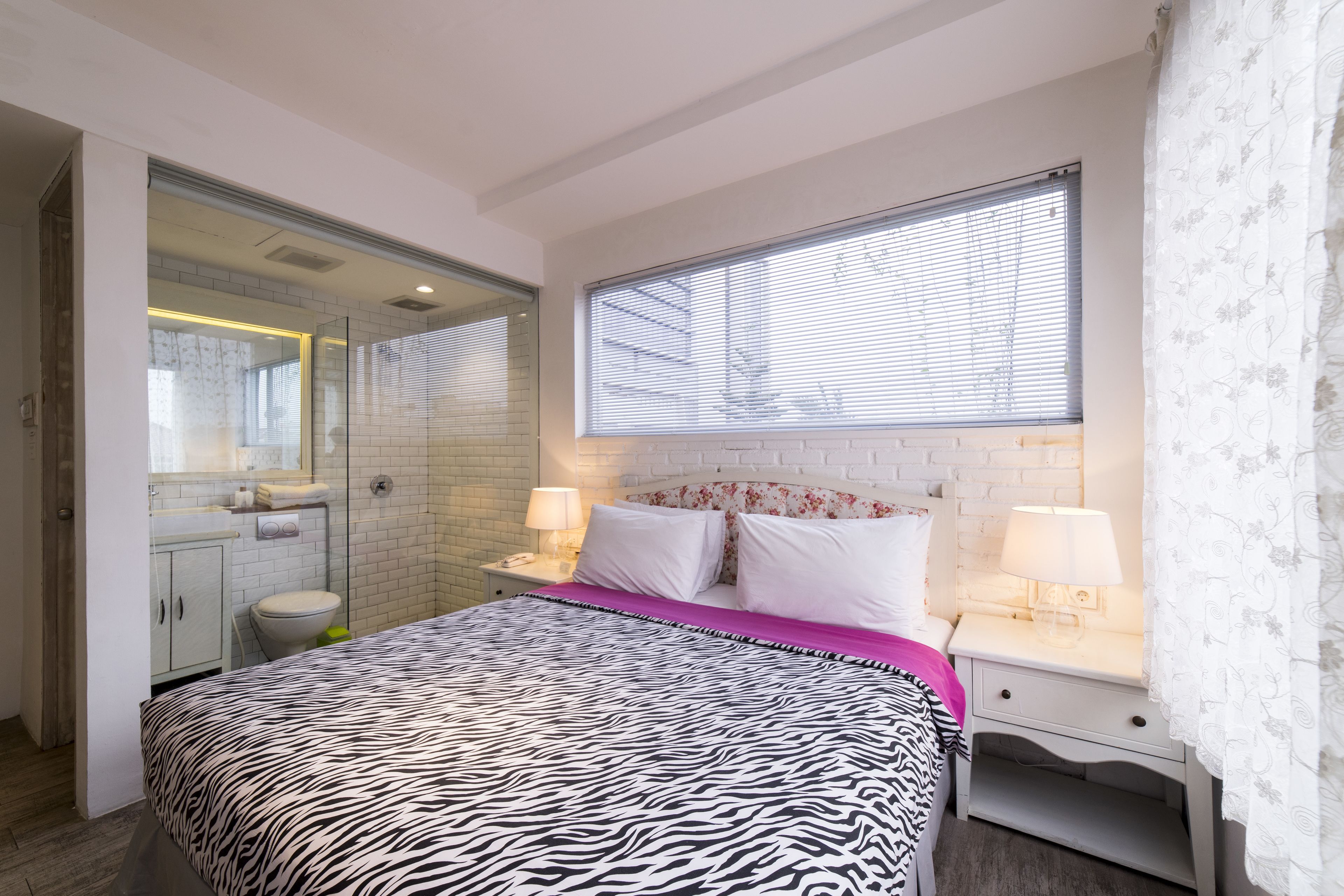 A bedroom with a zebra-patterned bedspread, a white brick wall, a large window with blinds, and a nightstand with a lamp.