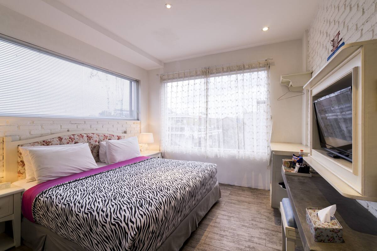A bedroom with a zebra-patterned bedspread, a television on a stand, and a window with sheer curtains.
