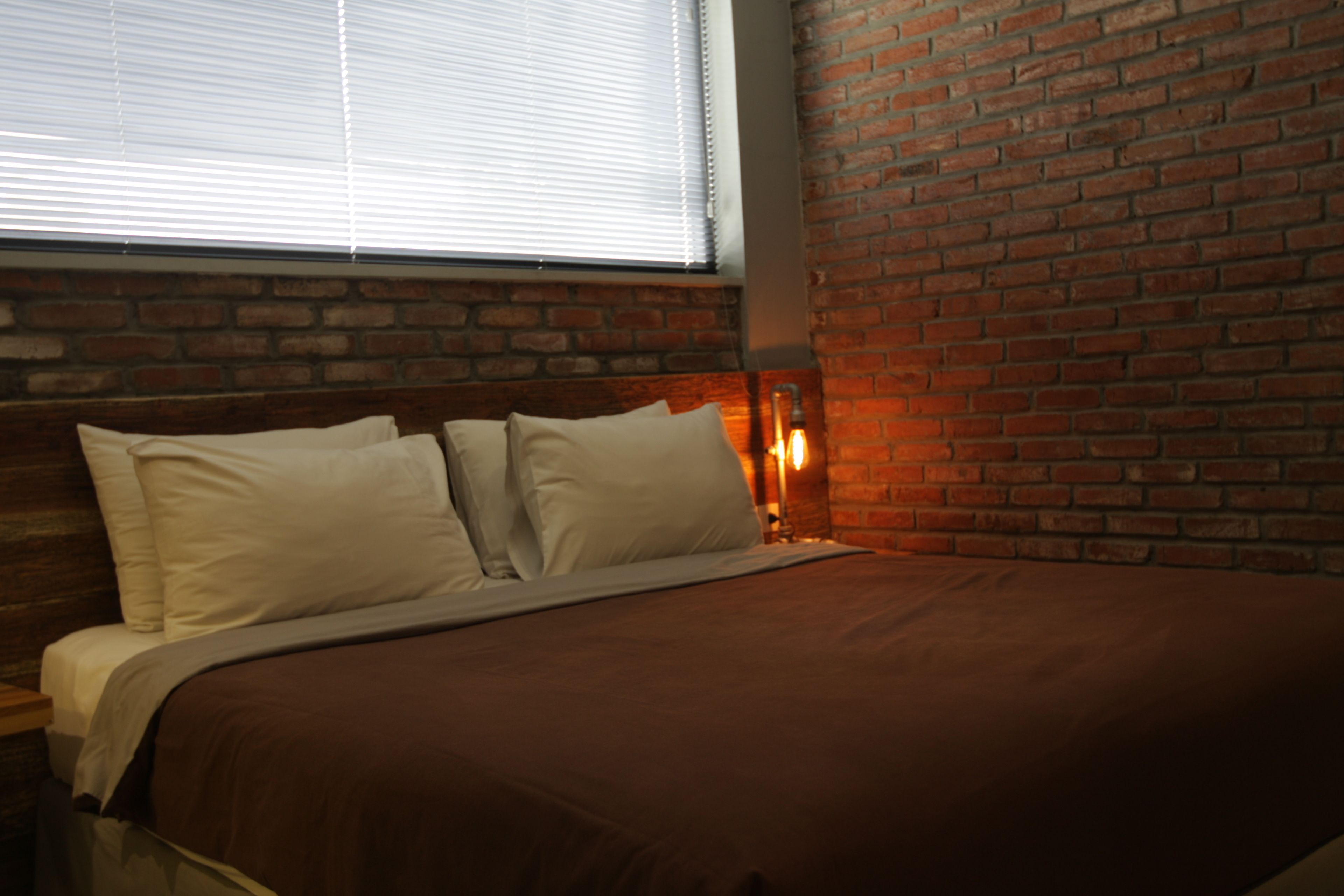 A bed with a brown bedspread and white pillows, a brick wall, and a window with blinds.
