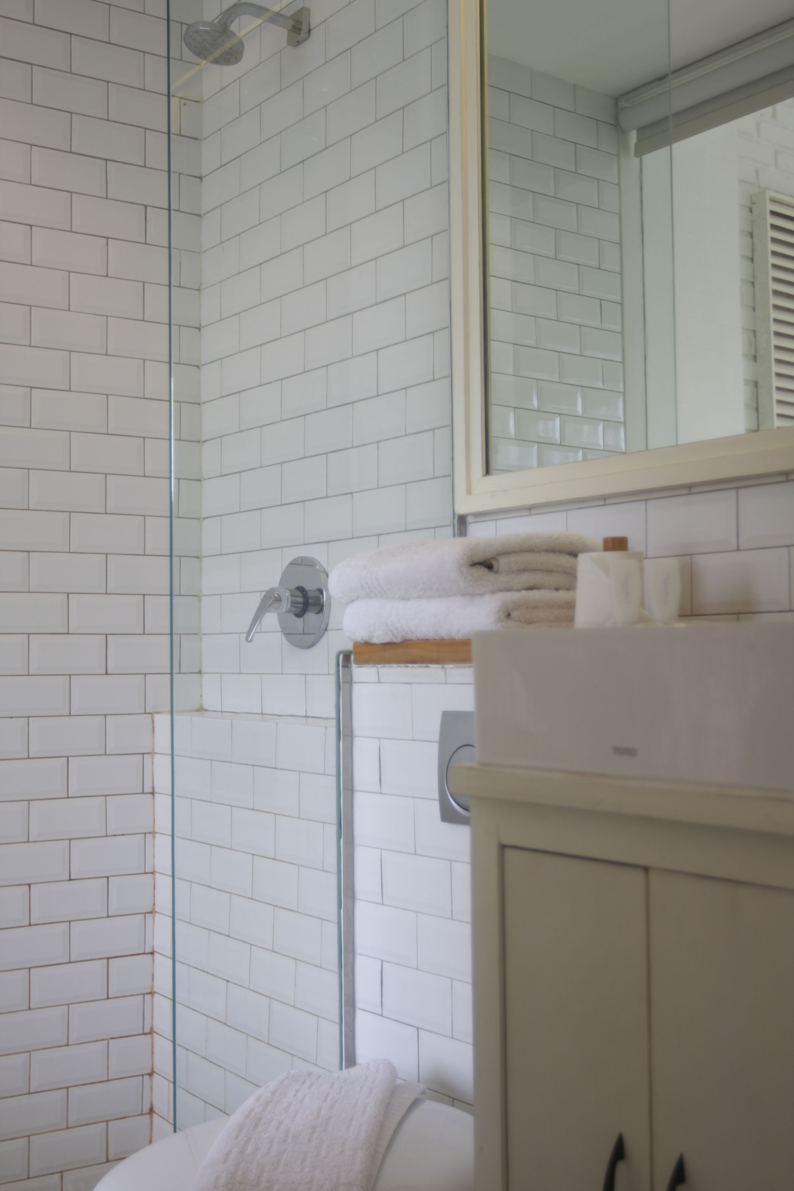 Ein Badezimmer mit weißen Fliesen, einer Duschkabine aus Glas, einem weißen Waschbecken und einem Spiegel darüber.