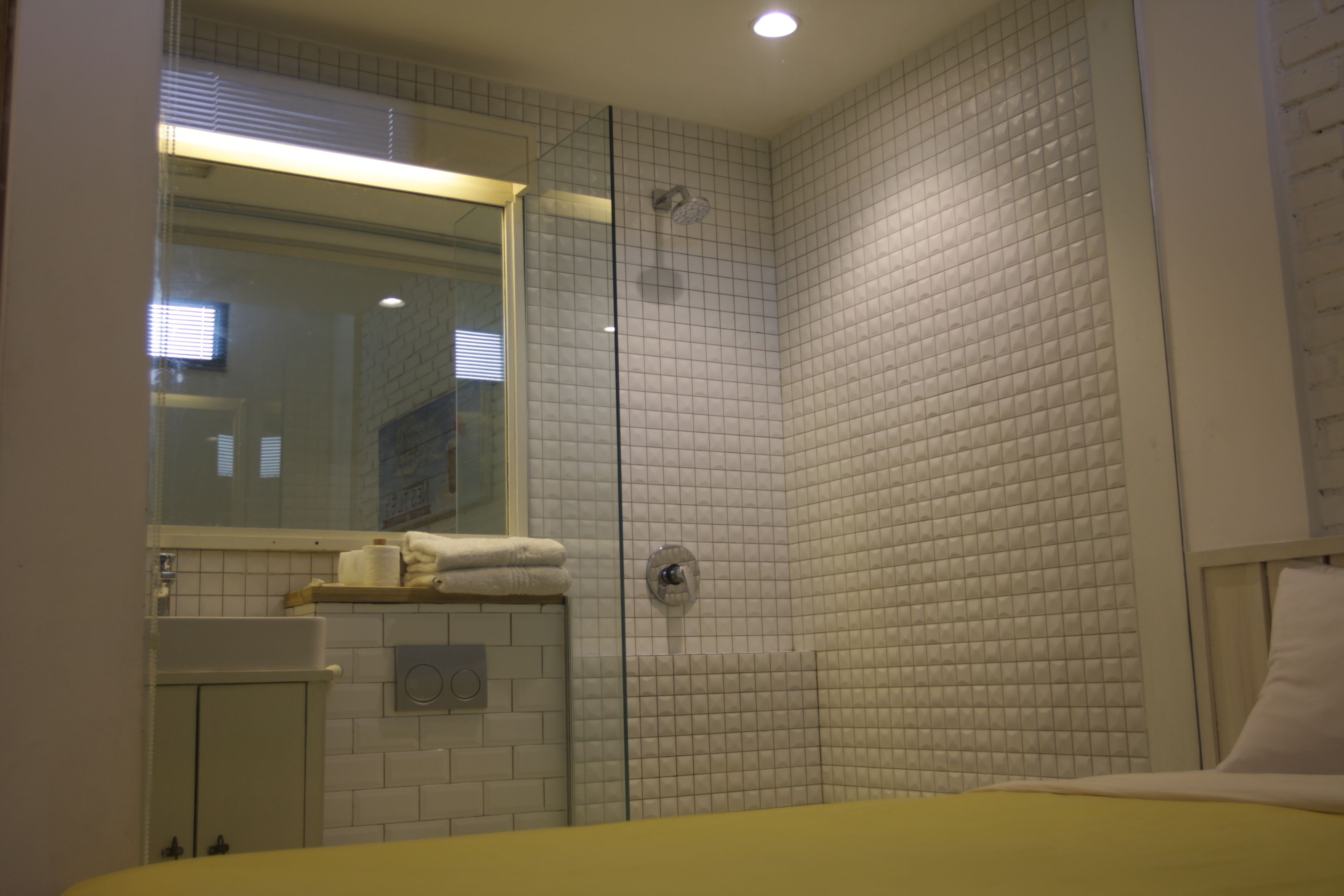 A bathroom with a glass shower enclosure, white tiled walls, a visible toilet, and a bed with a yellow cover.