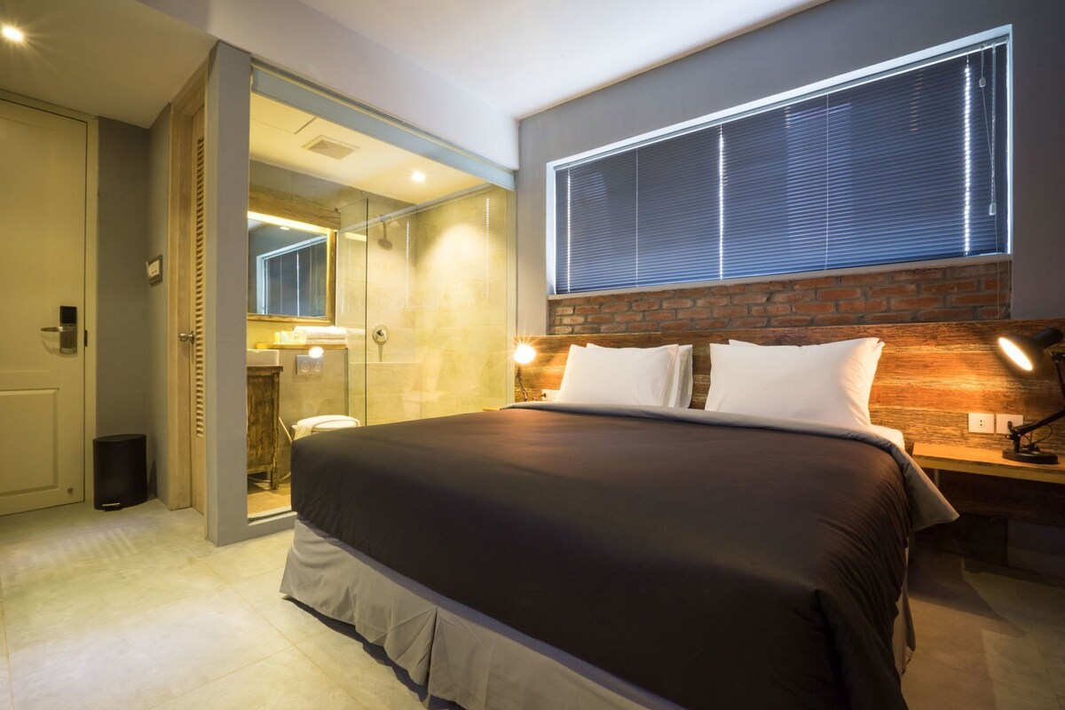 Une chambre d’hôtel moderne dotée d’un grand lit, d’une douche en verre, d’un mur décoratif en briques et d’une grande fenêtre avec stores.