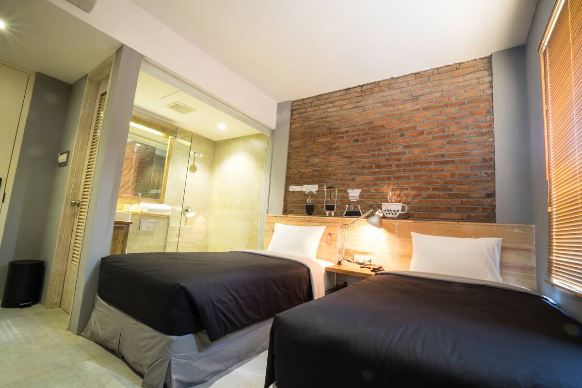A hotel room with two beds, a brick accent wall, a wooden headboard, a bedside table with a lamp, and a view of the bathroom through a glass door.