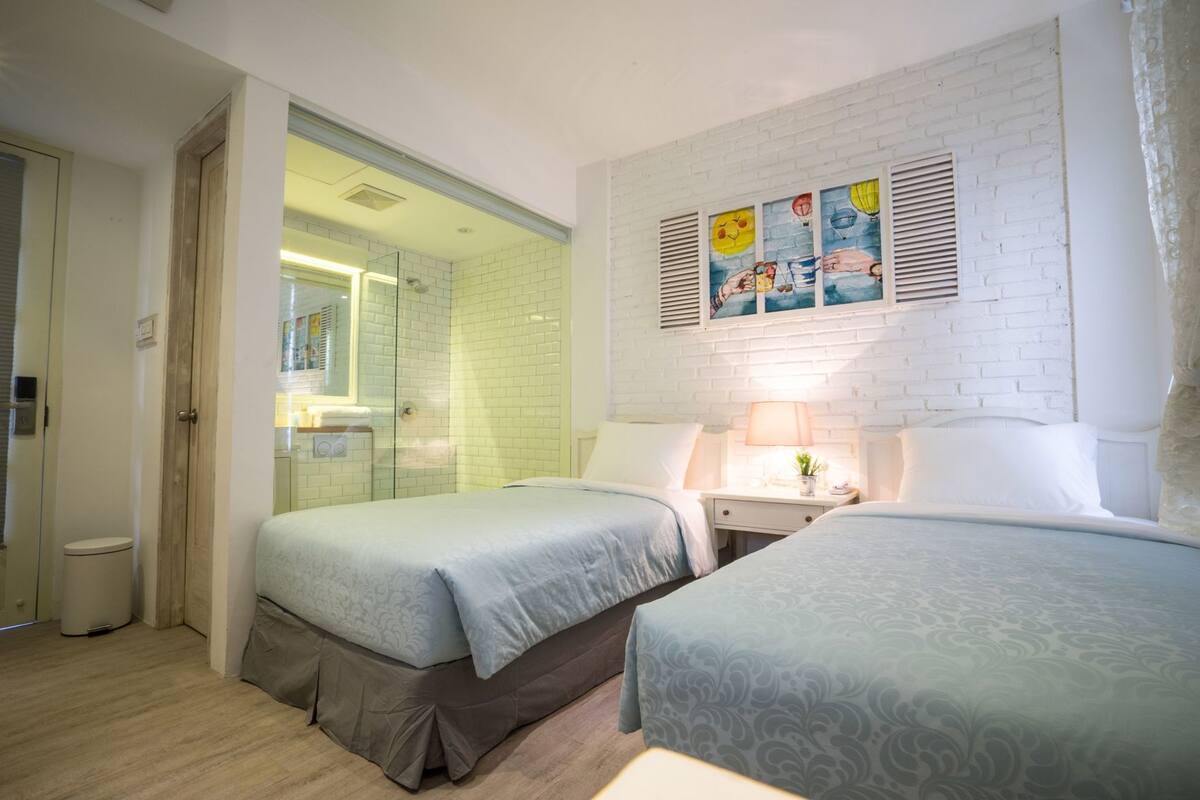 A hotel room with two beds, a bathroom, and a white brick wall with artwork.