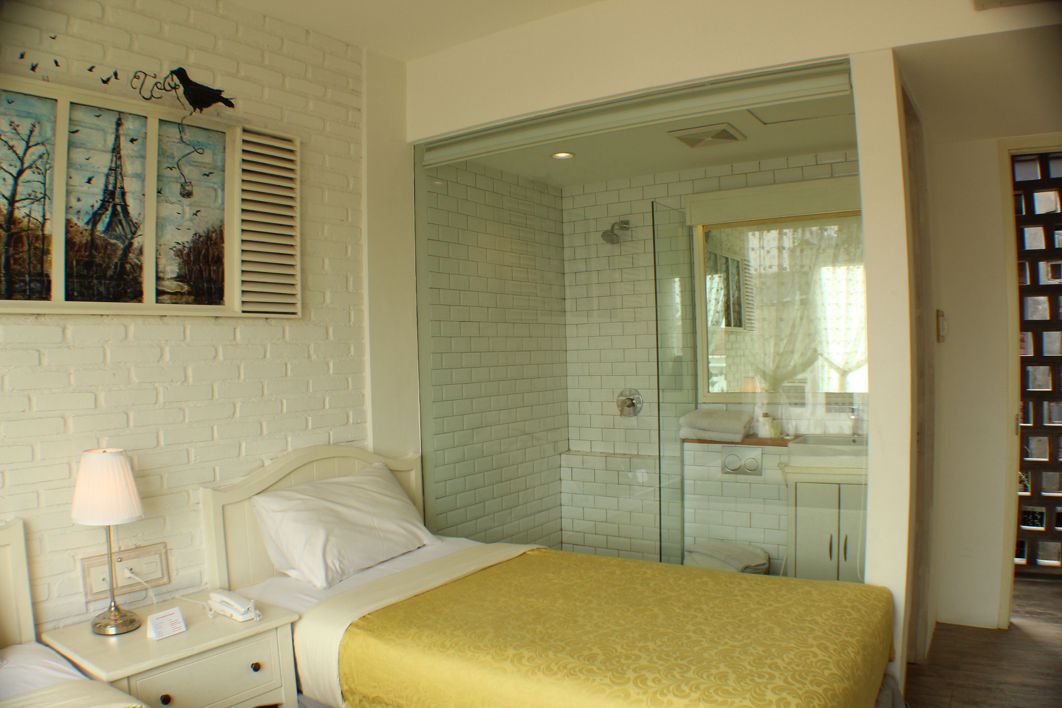 Quarto de hotel com cama, mesa de cabeceira, luminária, quadro, chuveiro e banheiro com espelho.