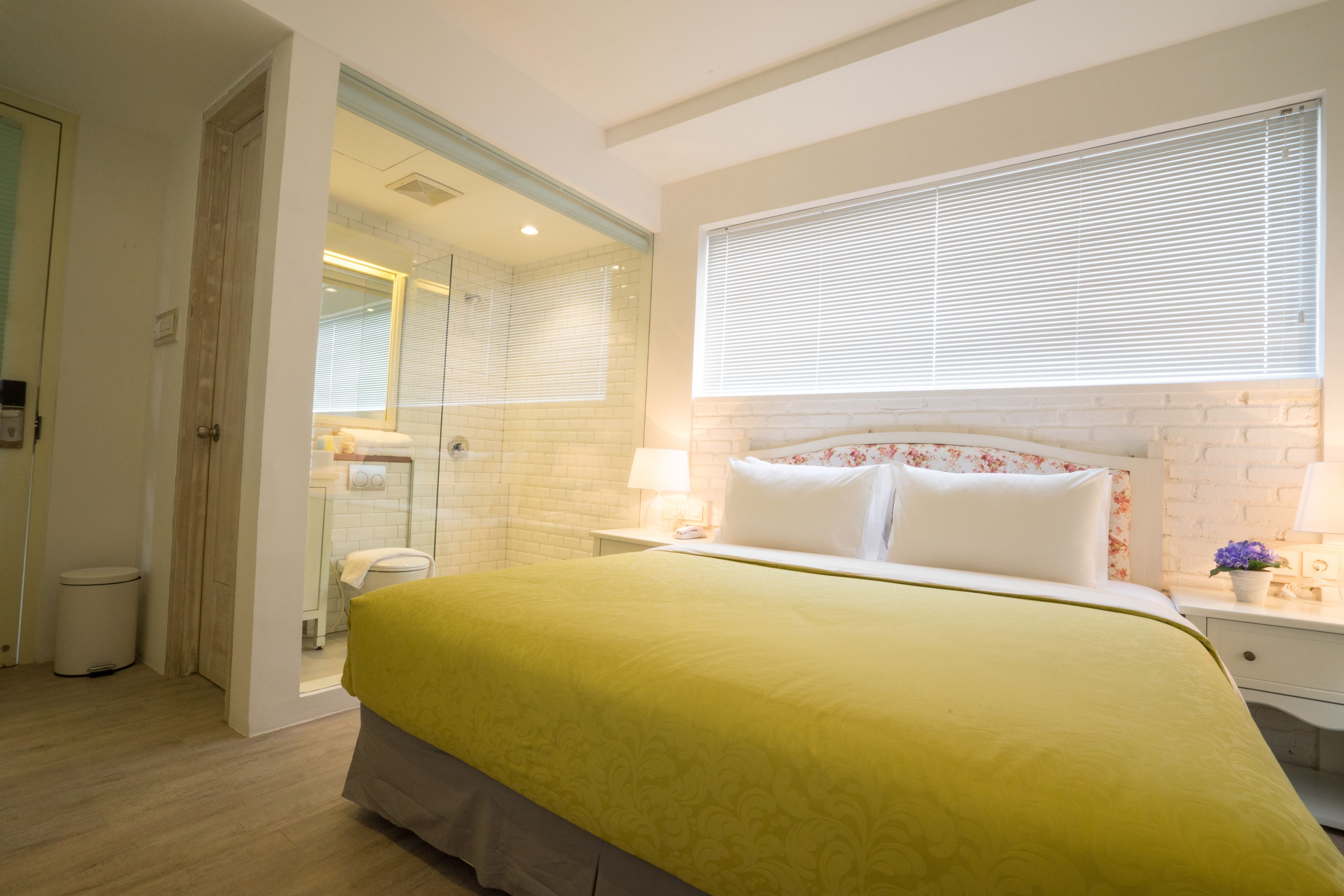 Un dormitorio con cama, ventana con persianas, mesita de noche con lámpara y baño visible a través de una puerta abierta.