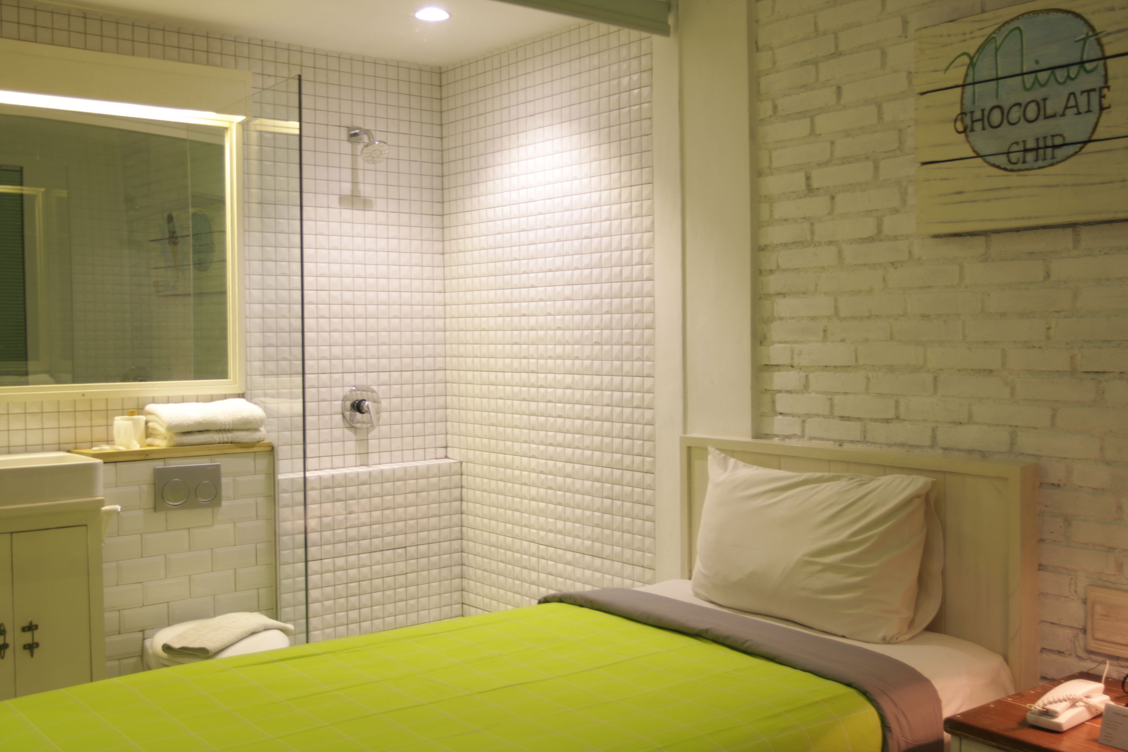 A hotel room with a green bed, a white wall with a 'Nitt Chocolate Chip' sign, a shower, and a small bedside table with a phone.