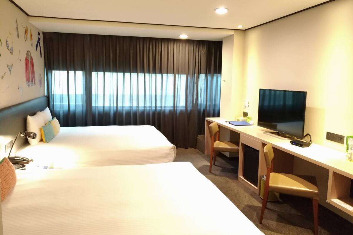 A hotel room with two beds, a desk with a TV, and a window with curtains.