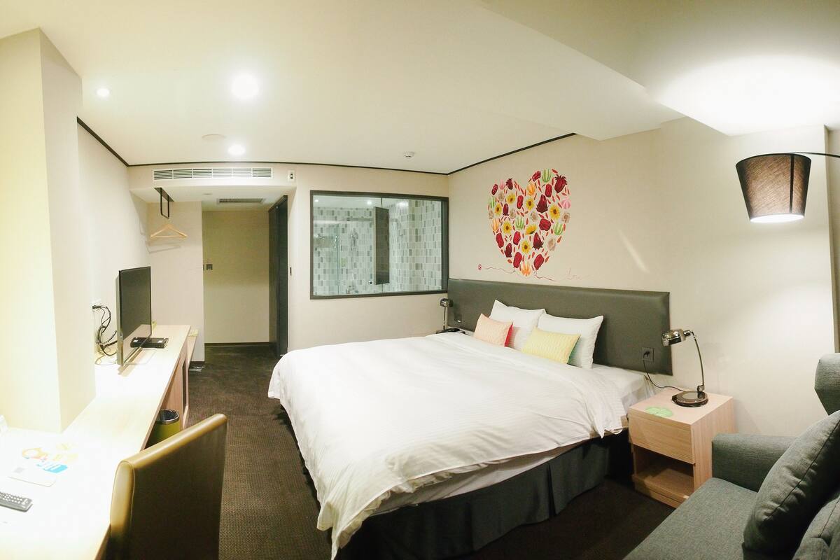 A hotel room with a bed, bedside table, lamp, and a desk with a chair.