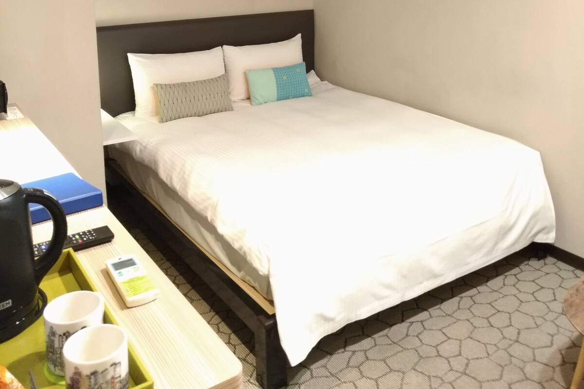 A hotel room with a bed, bedside table, and a desk with a coffee machine and mugs.
