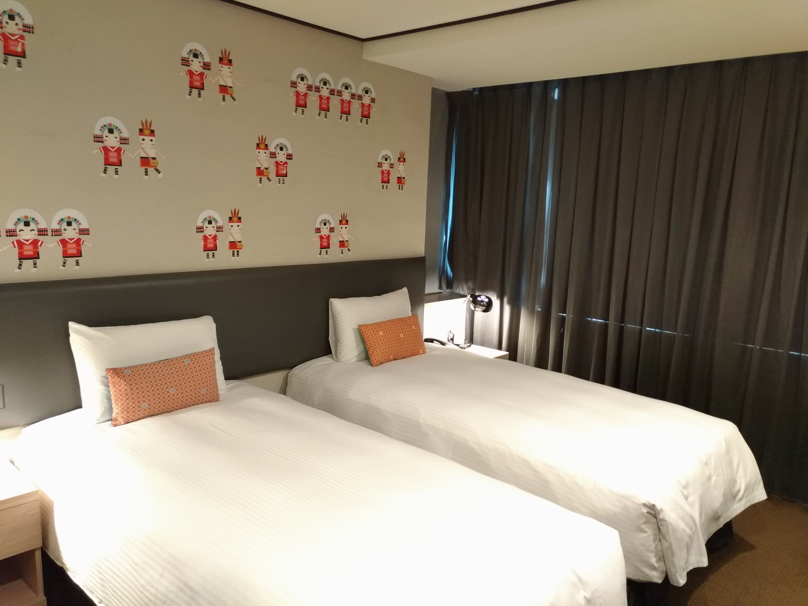 A hotel room with two beds, a large window with curtains, and a wall decorated with cultural figures.