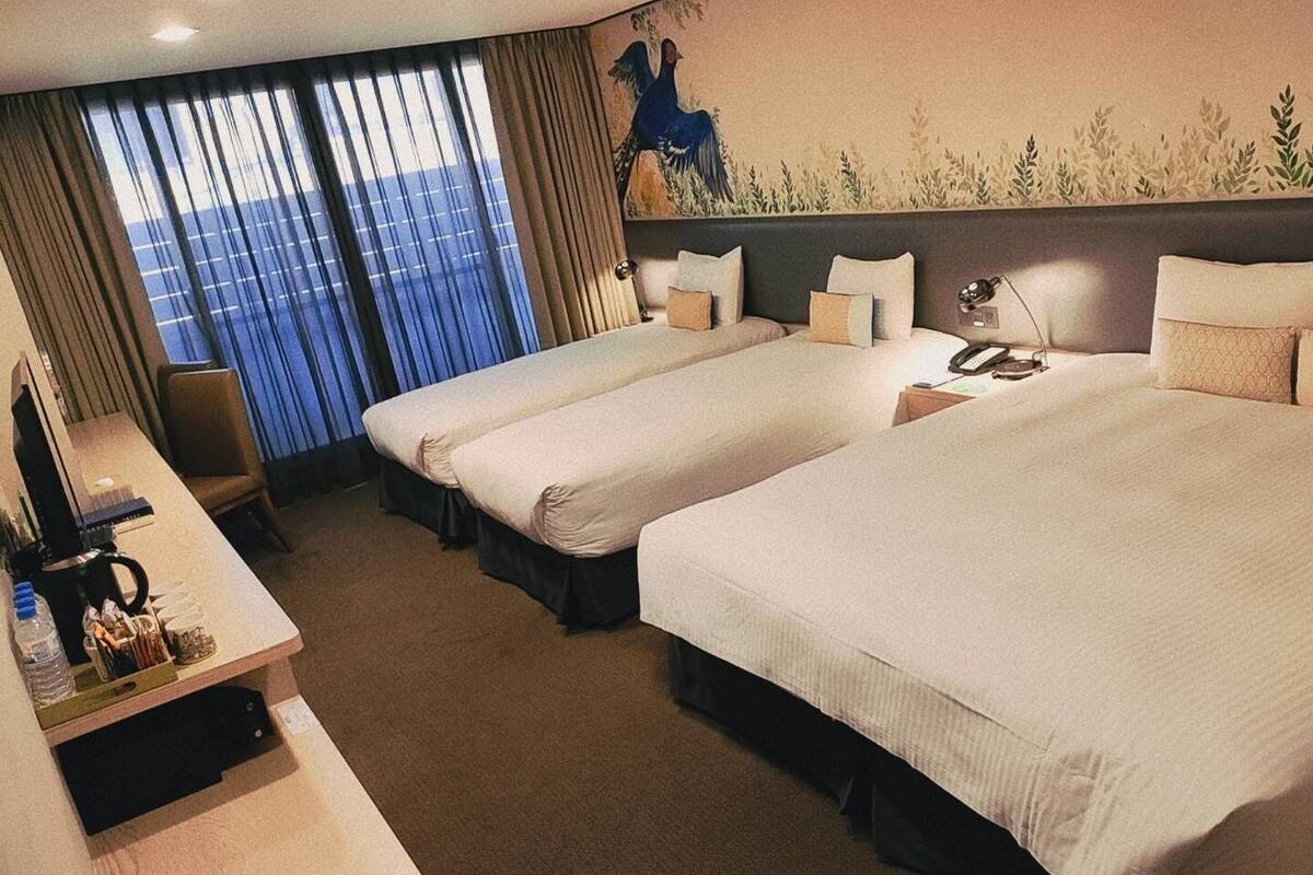 A hotel room with two beds, a desk with a coffee maker, and a large window with curtains.
