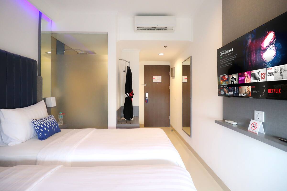 A hotel room with two beds, a TV wall with movie posters, a coat rack, and a bathroom visible through a glass partition.