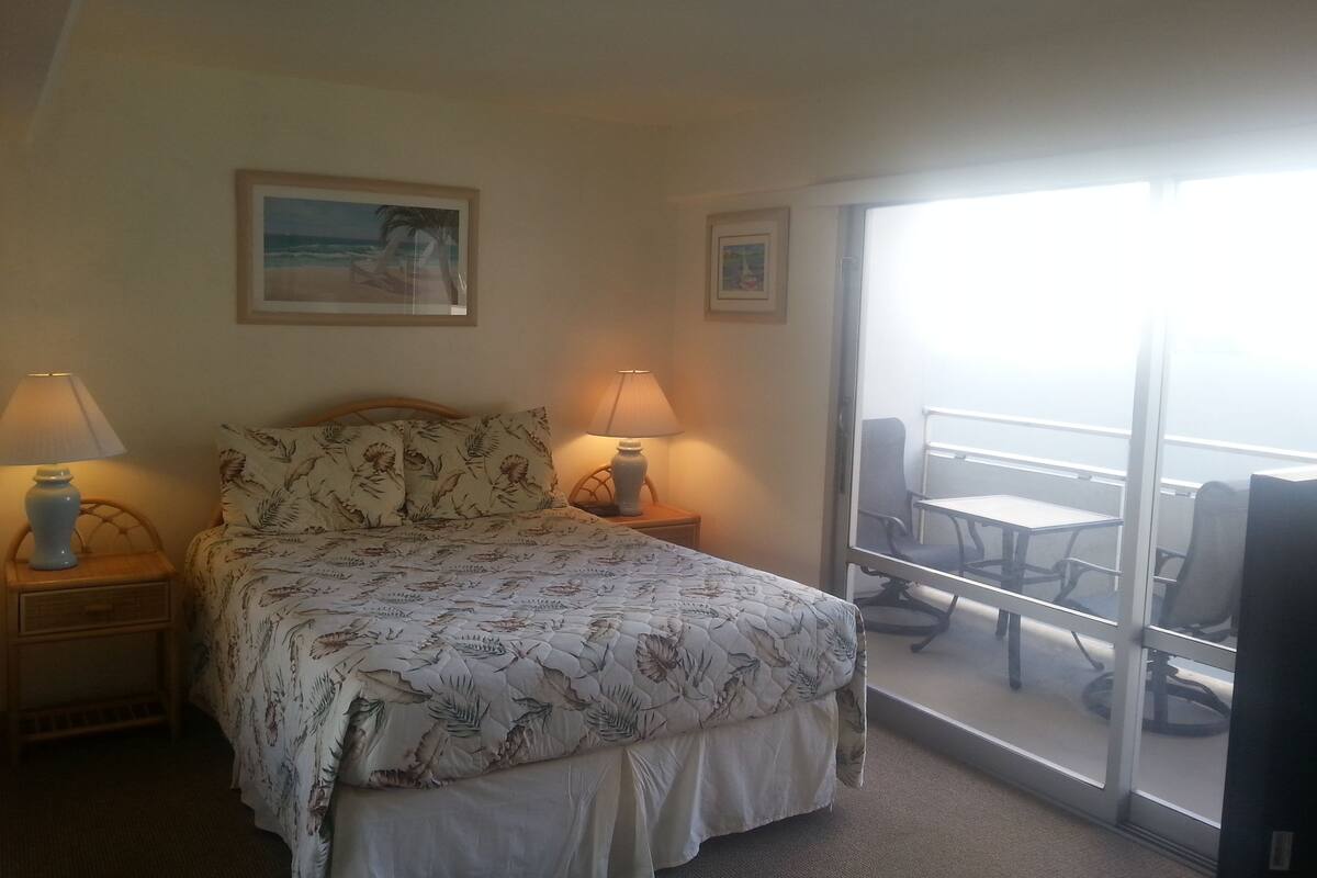 Une chambre d’hôtel avec un lit, deux lampes de chevet, un tableau au mur et un balcon avec des chaises.