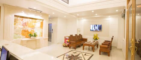 A spacious lobby with a marble reception desk, a wooden sofa set, a TV mounted on the wall, and a chandelier.