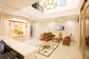 A spacious lobby with a marble reception desk, a wooden sofa set, a TV mounted on the wall, and a chandelier.