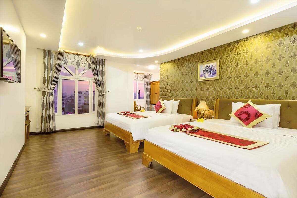 A hotel room with two beds, wooden flooring, a large window with curtains, a wall-mounted picture, and a ceiling light fixture.