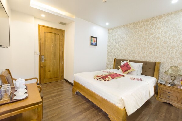 Una habitación de hotel con una cama de madera, una mesita de noche con lámpara, un escritorio de madera con tetera y tazas, y una puerta.